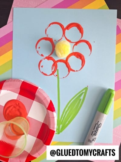 A children’s flower craft: red paint circles form petals, a yellow pom-pom is the center, and a green marker draws the stem on blue paper. Perfect for flower kids crafts, with a plastic cup, paper plate with paint, and marker nearby.