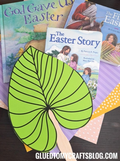 A large popsicle stick palm leaf craft sits on colorful paper next to three children's Easter books: God Gave Us Easter, The Easter Story, and The First Easter.