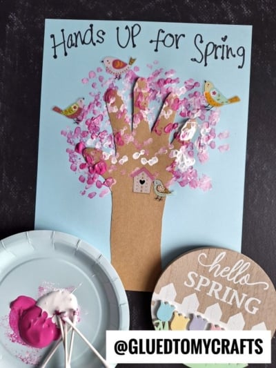 A spring-themed craft featuring a handprint tree with pink painted blossoms, paper birds, and a small birdhouse on blue paper—perfect for those seeking flower kids crafts or new flower craft ideas. Paint, cotton swabs, and a “hello SPRING” sign complete the scene.