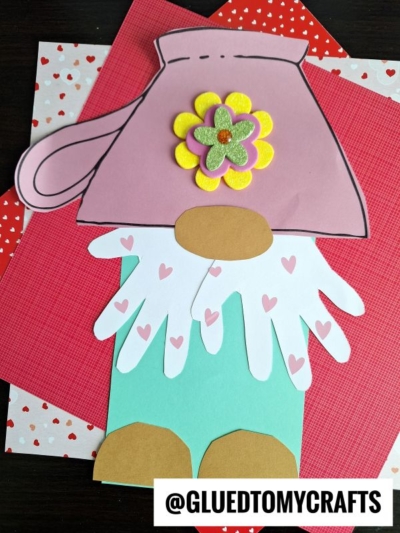 A teacup gnome made from colored paper shapes, featuring a pink hat with a flower, white handprint cutouts with hearts for the beard, teal pants, and brown shoes. The heart-patterned paper background adds extra charm.