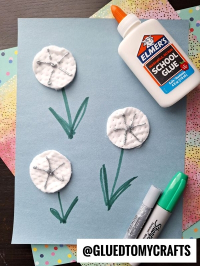 A fun flower kids crafts project: three dandelions made with cotton pads and glitter glue on blue paper. Stems and leaves are drawn in green marker, with Elmer's glue, a silver glitter pen, and two green markers nearby for creativity.