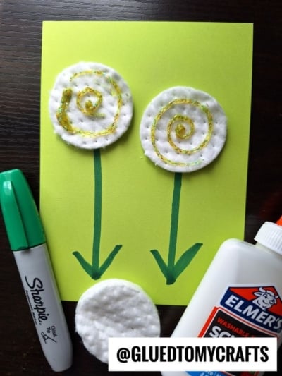 A handmade flower kids craft on green paper uses cotton pads for flower heads, gold glitter spirals, green marker stems, and two arrows. Nearby are a green marker, glue bottle, and loose cotton pad—great for creative flower craft ideas.