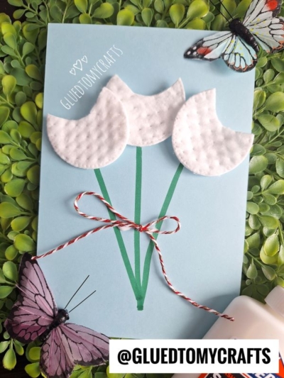 A handmade card featuring three white tulips made from cotton pads, perfect for flower craft ideas. With green stems, a red-white bow, leafy background, and butterfly decorations. The handle @GLUEDTOMYCRAFTS is visible.