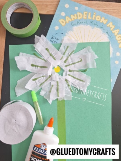A handmade dandelion craft with white tissue petals, green paper stem, and yellow center sits on green paper—perfect for flower craft ideas or flower kids crafts. Elmers glue, green tape, glitter, and a “Dandelion Magic” book are nearby.