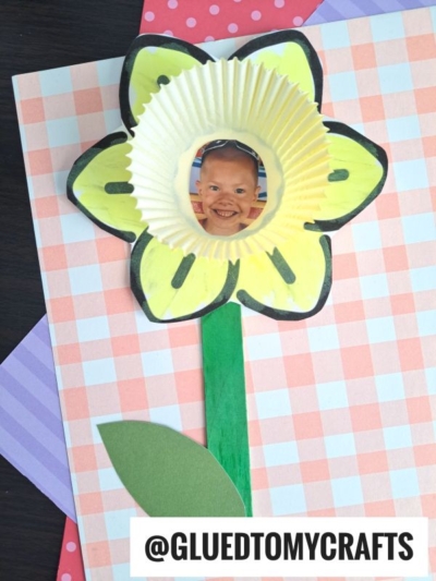 A handmade cupcake liner daffodil craft featuring a child’s smiling face at the center, with yellow petals, green paper stem and leaf, on a pink checkered background. @GLUEDTOMYCRAFTS is labeled at the bottom.