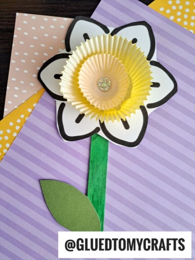 A handmade cupcake liner daffodil crafted from layered liners, black marker details, a glittery center, a green craft stick stem, and leaf, displayed on colorful patterned paper. @GLUEDTOMYCRAFTS appears at the bottom.
