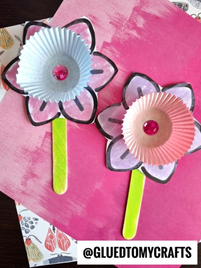 Two handmade cupcake liner daffodil crafts with drawn paper petals, pink gem centers, and green popsicle stick stems are displayed on a pink background. @GLUEDTOMYCRAFTS is written at the bottom.
