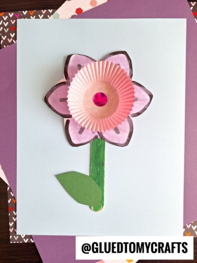 A handmade cupcake liner daffodil crafted from pink cupcake liners, featuring a red gem center, green paper stem and leaf, on a light blue background. The username @gluedtomycrafts appears at the bottom.