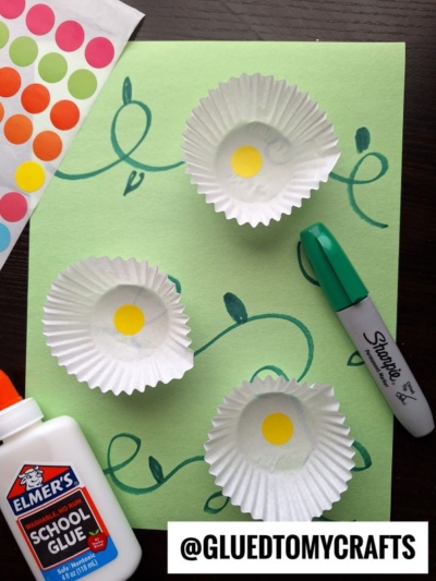 A craft setup with green paper loops, three cupcake liners each with a yellow dot in the center, a green Sharpie, Elmers glue, and colorful dot stickers—perfect for flower kids crafts. The handle @GLUEDTOMYCRAFTS is displayed.