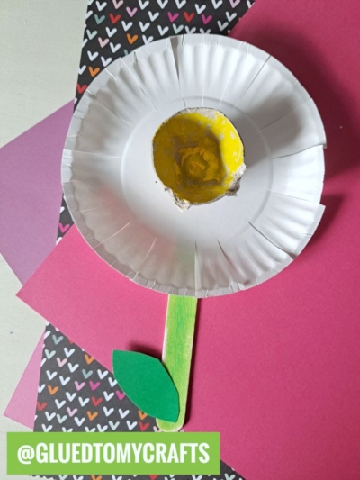 A paper craft flower made from a white cupcake liner with a yellow center and green popsicle stick stem, placed on pink and black heart-patterned paper. Perfect for flower kids crafts or anyone looking for creative flower craft ideas. @GLUEDTOMYCRAFTS.