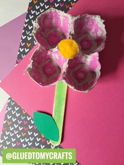 A pink flower craft made from an egg carton section with a yellow pom-pom center, green popsicle stick stem, and paper leaf—one of the cutest flower kids crafts—displayed on colorful paper backgrounds.