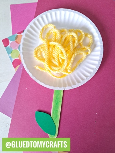 A paper plate with yellow yarn loops forms a dandelion, paired with a green craft stick stem and paper leaf—fun flower kids crafts or flower craft ideas. Displayed on red and multi-colored papers. @GLUEDTOMYCRAFTS at the bottom.