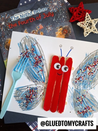 A Fourth of July fork painted butterfly craft with a red Popsicle stick body, googly eyes, and blue and red glitter wings shaped using a plastic fork, displayed on The Night Before the Fourth of July book and black paper.