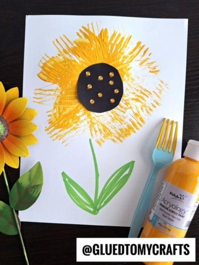 A sunflower painting made with yellow paint, a green stem and leaves, and a black center with gold dots—perfect for flower kids crafts. A blue fork, fake sunflower, and yellow paint bottle sit next to the artwork. @GLUEDTOMYCRAFTS is written at the bottom.