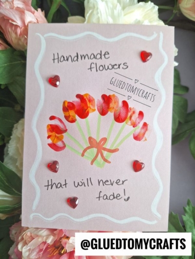 A handmade pink greeting card decorated with painted red and orange flowers, small red heart gems, and “Handmade flowers that will never fade!”—perfect for flower craft ideas. Surrounded by real blooms. @GLUEDTOMYCRAFTS appears at the bottom.