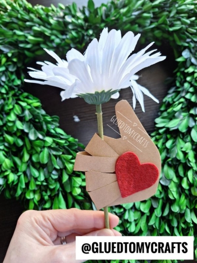 A hand holds a white artificial daisy with a paper cutout of a hand wrapped around the stem, decorated with a red felt heart—perfect for flower kids crafts. Green wreath leaves are blurred in the background. Text: @GLUEDTOMYCRAFTS.