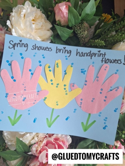 A blue paper displays three colorful handprint cutouts as flowers, decorated with blue dots and green stems—perfect for anyone seeking flower craft ideas. The text reads Spring showers bring handprint flowers! Pink flowers surround the artwork.