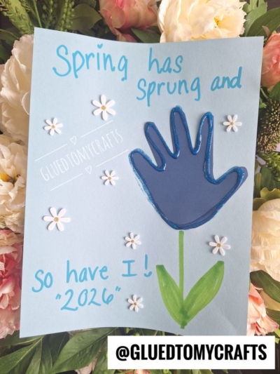 A light blue card features a dark blue painted handprint as a flower, with green stem and leaves, surrounded by white paper flowers—a charming pick for flower kids crafts. Text reads: “Spring has sprung and so have I! 2026.” @GLUEDTOMYCRAFTS is at the bottom.