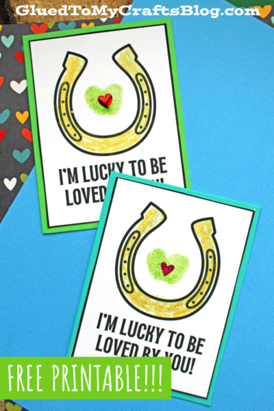 Two printable Fingerprint St. Patrick's Day cards feature a gold horseshoe, green heart, and red heart. Below the horseshoe is the message: “I’M LUCKY TO BE LOVED BY YOU!” Cards are displayed on a blue, heart-patterned background.
