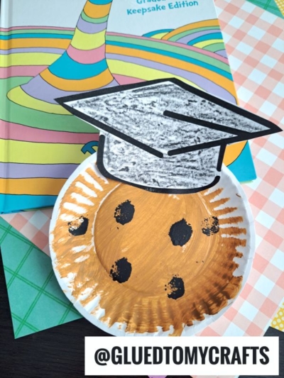 A paper plate smart cookie is painted gold with black spots, topped with a large paper graduation cap for a fun graduation-themed craft. Colorful books appear in the background, and @GLUEDTOMYCRAFTS is displayed at the bottom.