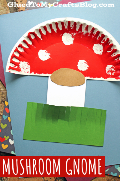 A children’s craft featuring a whimsical mushroom gnome made from a painted paper plate, brown oval paper, green fringed grass, and a blue paper background. The caption reads, MUSHROOM GNOME.