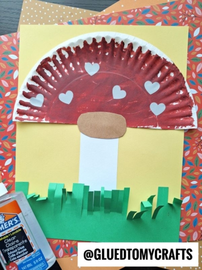 A children's mushroom gnome craft made with a painted paper plate cap, brown paper stem and base, heart-shaped spots, and green paper grass on a yellow background. Elmer’s glue and colored paper are nearby.