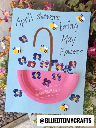 A blue craft paper with “April showers bring May flowers” written on it, decorated with painted flowers, bee stickers, and a pink paper umbrella—perfect for flower kids crafts inspiration. Handle drawn with brown marker. @GLUEDTOMYCRAFTS tag at the bottom.