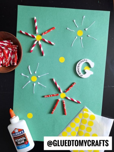 Green paper with white glue, red-striped straws arranged like flowers, and yellow dot stickers—perfect for easy flower kids crafts. An Elmer’s glue bottle, a dish with straws, yellow stickers, and a logo card sit nearby. Text: “@GLUEDTOMYCRAFTS.”.