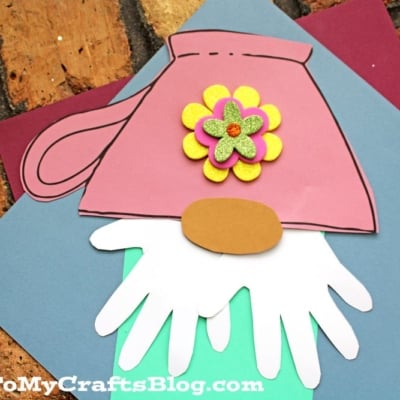 A colorful paper craft teacup gnome features a pink teacup hat, white beard, brown nose and shoes, mint green body, and a decorative yellow and purple flower on the hat.