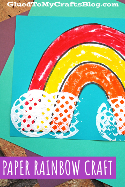 Colorful child's paper craft of a potato masher rainbow painted on blue paper, with clouds made from textured white paint. The image includes the text “GluedToMyCraftsBlog.com” and “PAPER RAINBOW CRAFT” at the bottom.
