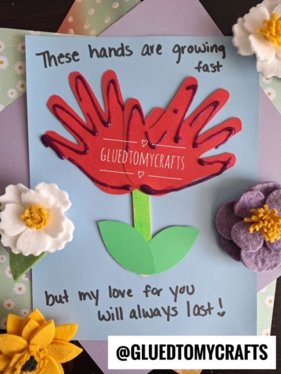 A colorful handmade card features a red handprint flower with green stem and leaves, surrounded by felt flower decorations—an adorable example of flower craft ideas. Text reads: These hands are growing fast but my love for you will always last!.