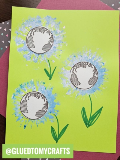 A handmade craft on yellow paper features three Earth Day flowers—dandelions with Earth globes as centers. The fluffy parts are blue and white, with green stems and leaves drawn. @GLUEDTOMYCRAFTS is written in white on green.