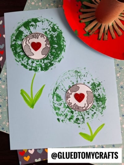 A blue paper displays two Earth Day flowers painted with green paint using a cardboard tube. Each dandelion has a white circle with a red heart and black designs in the center. @GLUEDTOMYCRAFTS is written at the bottom.