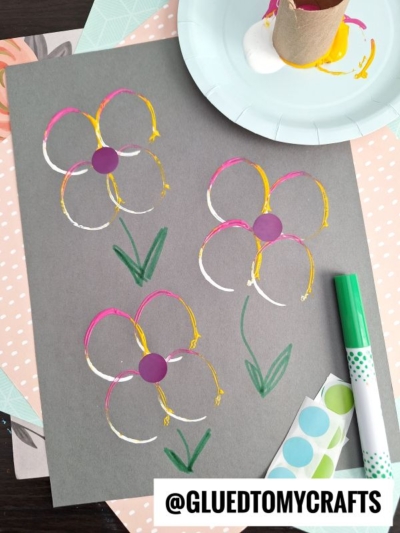 This flower kids crafts project features three colorful flowers stamped on gray paper with a cardboard tube and paint, green stems drawn in marker, and purple circle stickers at the centers. Art supplies are visible around the paper.