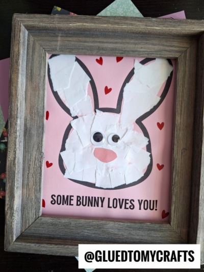 A framed, handmade torn paper bunny art on pink paper with red hearts; the bunny is made from torn white paper, has googly eyes, and the text SOME BUNNY LOVES YOU! appears at the bottom. @GLUEDTOMYCRAFTS is in the corner.