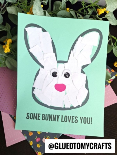 A handmade card featuring a torn paper bunny face with googly eyes and a pink nose on green paper. Text below reads SOME BUNNY LOVES YOU! Leaves and flowers decorate the background.