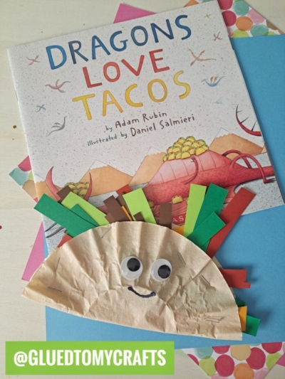 A children’s book titled Dragons Love Tacos sits on colorful papers, topped with a coffee filter taco craft made from a brown coffee filter, googly eyes, and colored paper strips. @GLUEDTOMYCRAFTS is written at the bottom.