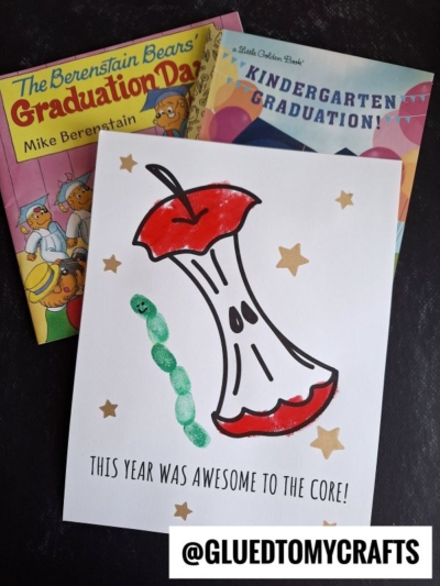 A card with a red apple core drawing, a fingerprint worm craft green caterpillar, and gold stars reads “This year was awesome to the core!” Two graduation-themed children’s books are in the background. @GLUEDTOMYCRAFTS is at the bottom.