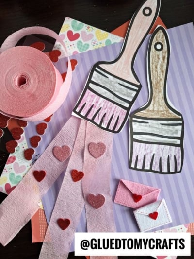 Two paper paint brush craft projects with pink and brown handles rest on striped and heart-patterned paper. Pink crepe streamers with red hearts and small paper envelopes are nearby. @GLUETOMYCRAFTS text appears at the bottom.