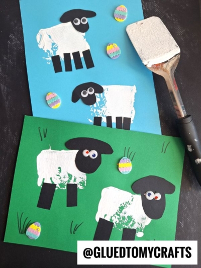 Two sheep crafts made from white paint handprints, black paper, and googly eyes are displayed on blue and green paper with colorful paper Easter eggs. This fun spatula stamped crafts project uses a spatula with white paint, seen resting nearby. @GLUEDTOMYCRAFTS is written at the bottom.
