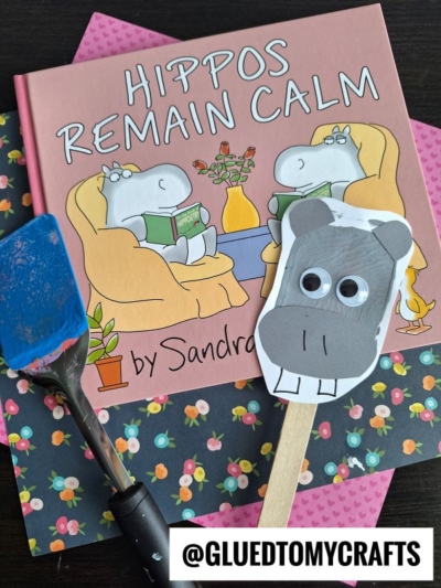 A children’s book titled Hippos Remain Calm is shown with a blue spatula, floral paper, and a hippo craft stick with googly eyes—perfect inspiration for spatula stamped crafts. The @GLUEDTOMYCRAFTS handle appears at the bottom of the image.