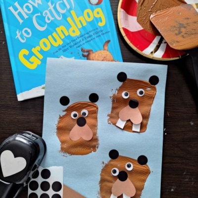 A children’s book titled How to Catch a Groundhog, a craft sheet with four painted groundhogs, spatula stamped crafts, black dot stickers, a punch tool, brown paint on a plate, and the tag @GLUEDTOMYCRAFTS displayed on a table.