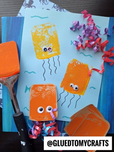 A blue paper showcases three painted orange sponge jellyfish with googly eyes and curled paper tentacles. This fun spatula stamped crafts idea includes a paint spatula, orange sponges, and the @gluedtomycrafts handle.