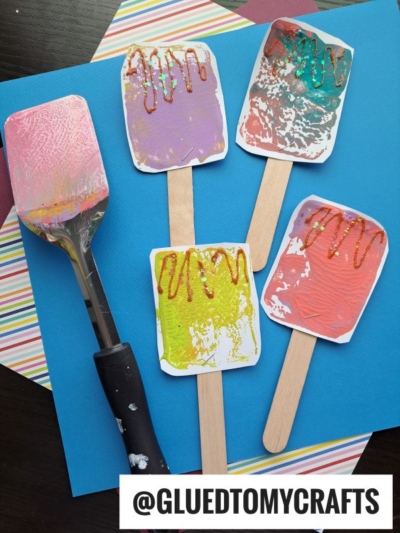 Four colorful popsicle crafts made of painted paper and wooden sticks are displayed on a blue surface, featuring glitter details. A paint-covered spatula hints at fun spatula stamped crafts, with striped paper partially visible underneath.
