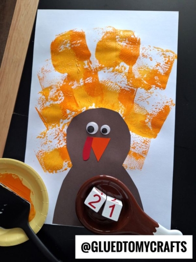 A turkey craft made from brown, red, and orange paper, with googly eyes and an orange beak. Yellow and orange paint marks, created using spatula stamped crafts techniques, form feathers behind the turkey. Art supplies and a @GLUEDTOMYCRAFTS label are visible.