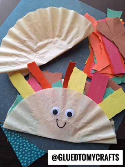 A paper turkey craft made from a folded coffee filter taco, decorated with googly eyes, a drawn smile, and colorful paper strips for feathers. Torn paper pieces are scattered nearby. @gluedtomycrafts is written at the bottom.