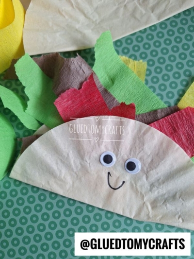 A coffee filter taco craft with googly eyes, a smiling face, and colored crepe paper for lettuce, cheese, and tomatoes sits on a green patterned surface. @GLUEDTOMYCRAFTS is written at the bottom.