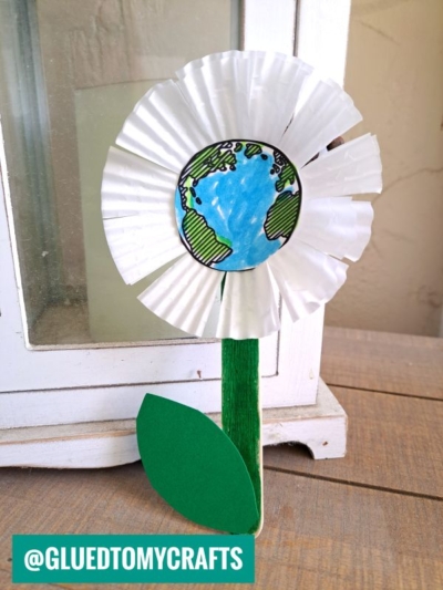 A handmade cupcake liner earth day flower features white paper petals, a globe drawing as the center, a green popsicle stick stem, and a green paper leaf, resting on a wooden surface. A watermark reads @GLUEDTOMYCRAFTS in the corner.