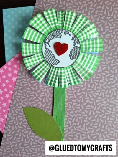 Handmade cupcake liner Earth Day flower craft with a globe and red heart center, surrounded by green and white petals, green stem, and leaf on a patterned background. Text reads @GLUEDTOMYCRAFTS.
