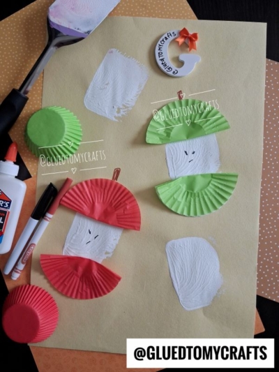 Craft supplies and green and red cupcake liners are arranged on paper, with some liners cut and glued to resemble an apple core. Glue, a black marker, white paint, and a paint spatula—perfect for spatula stamped crafts—are nearby. @GLUEDTOMYCRAFTS is labeled.